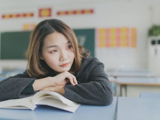 A woman sits with her arms crossed over an open book while in a classroom setting.